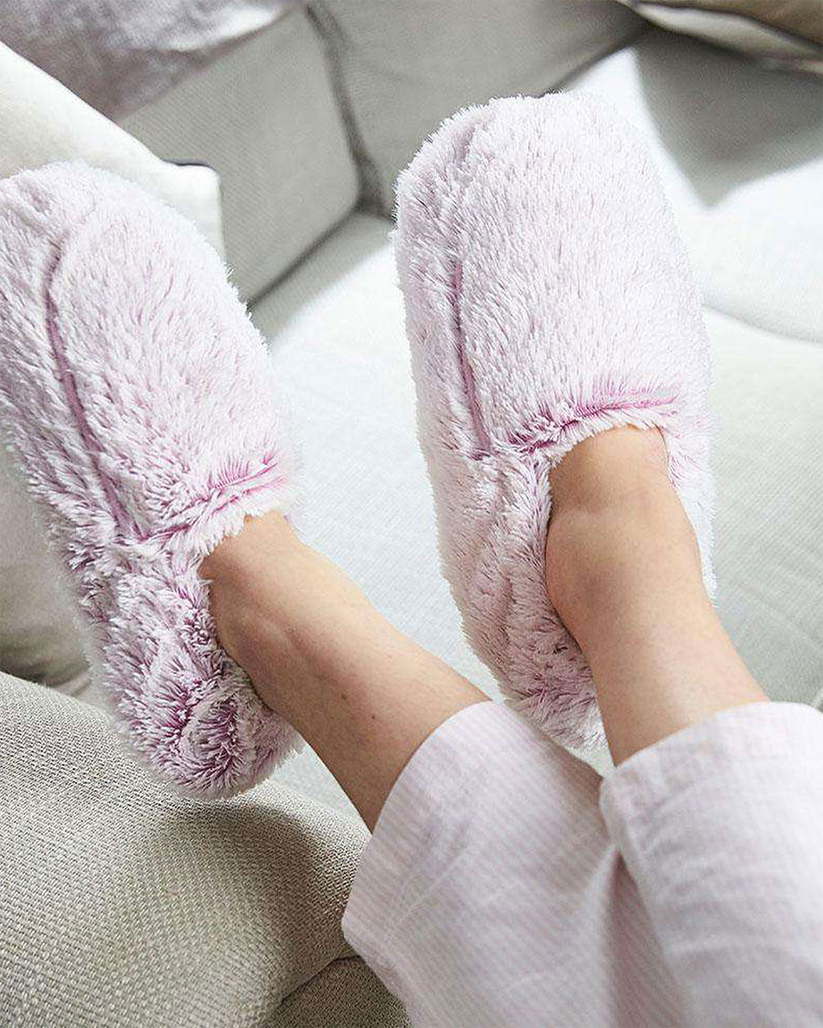 Person's legs resting on a gray sofa, wearing a pair of fuzzy light lavendar slippers with warmable inserts