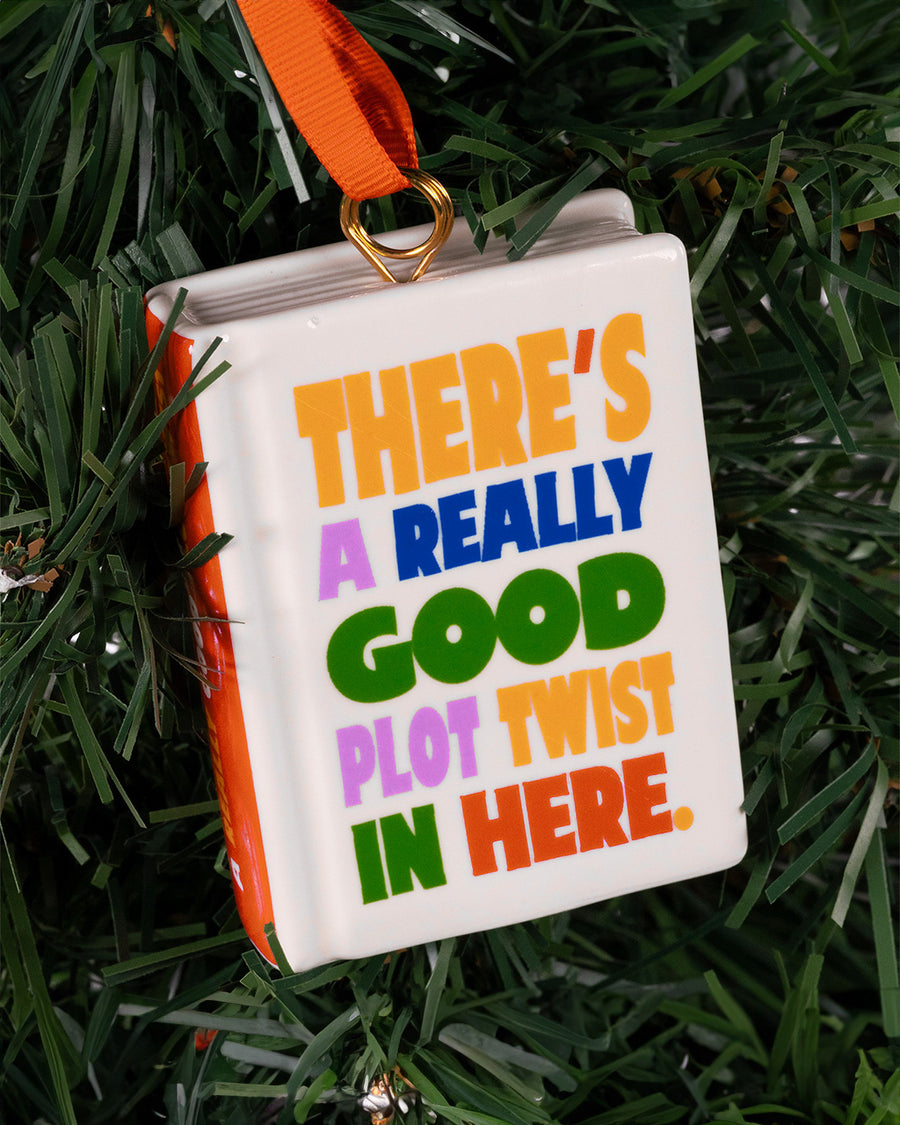 ceramic book shaped ornament with 'there's a really good plot twist in here' on the front and back hanging in a tree