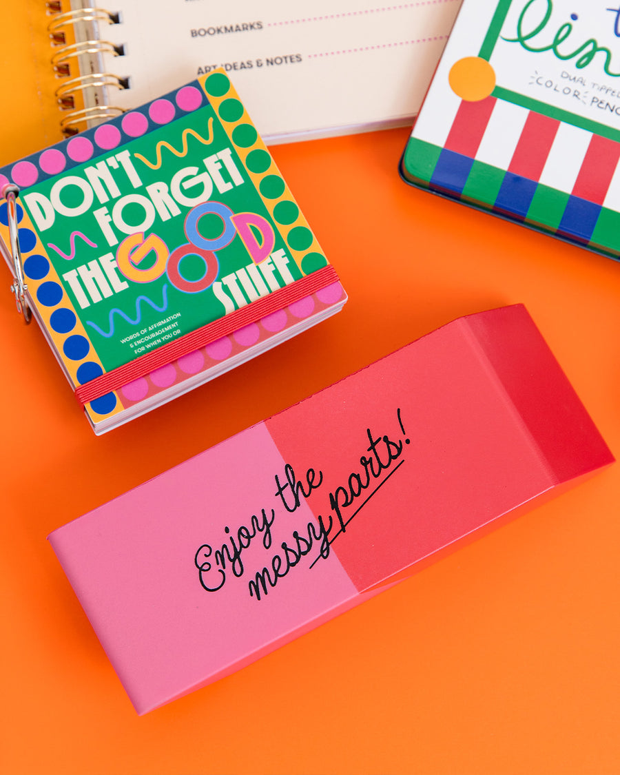 pink and red eraser shaped de-stress ball with black 'enjoy the messy parts' across the front on a desk