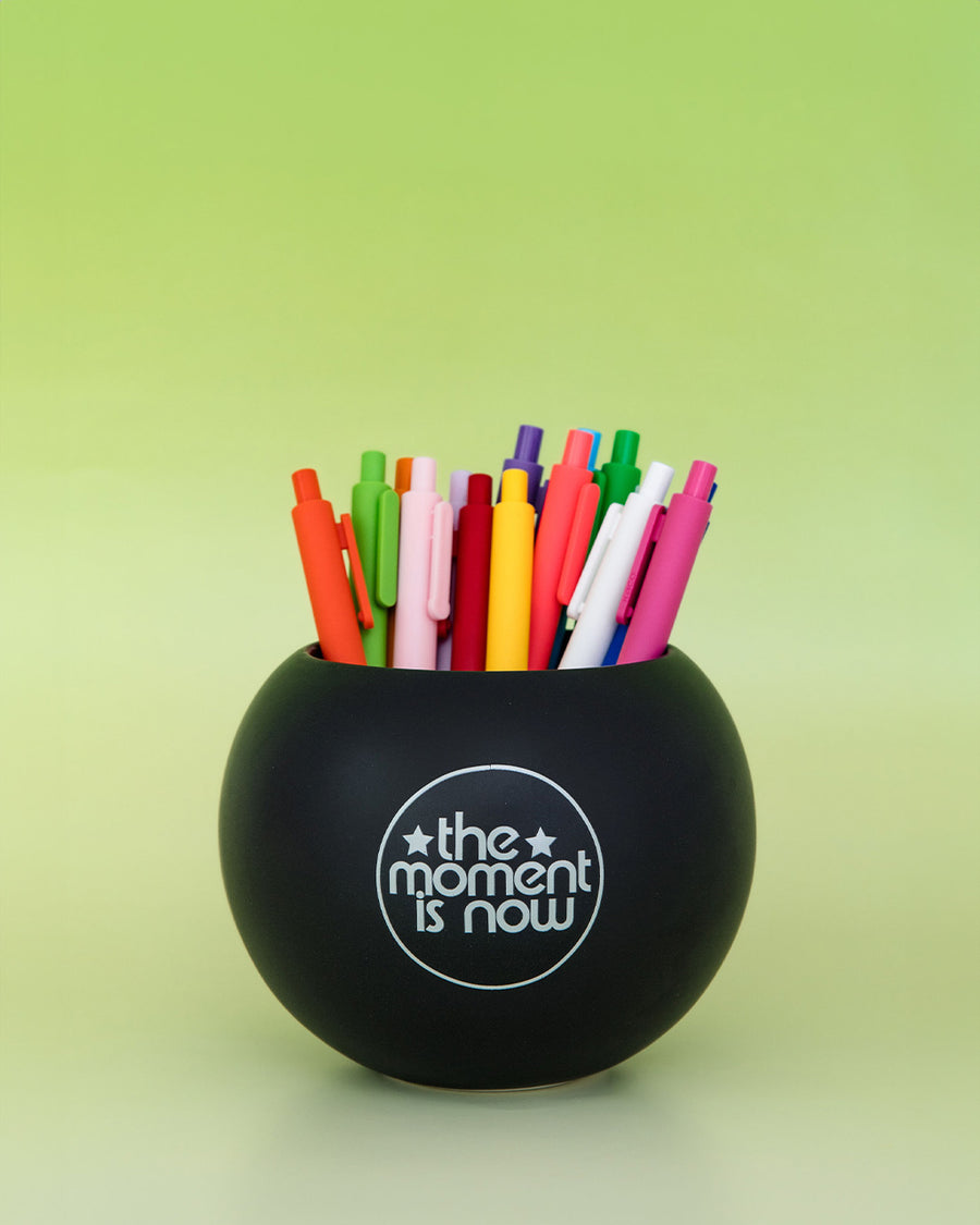 black magic 8 ball shaped vase with pens inside and 'the moment is now' on the back