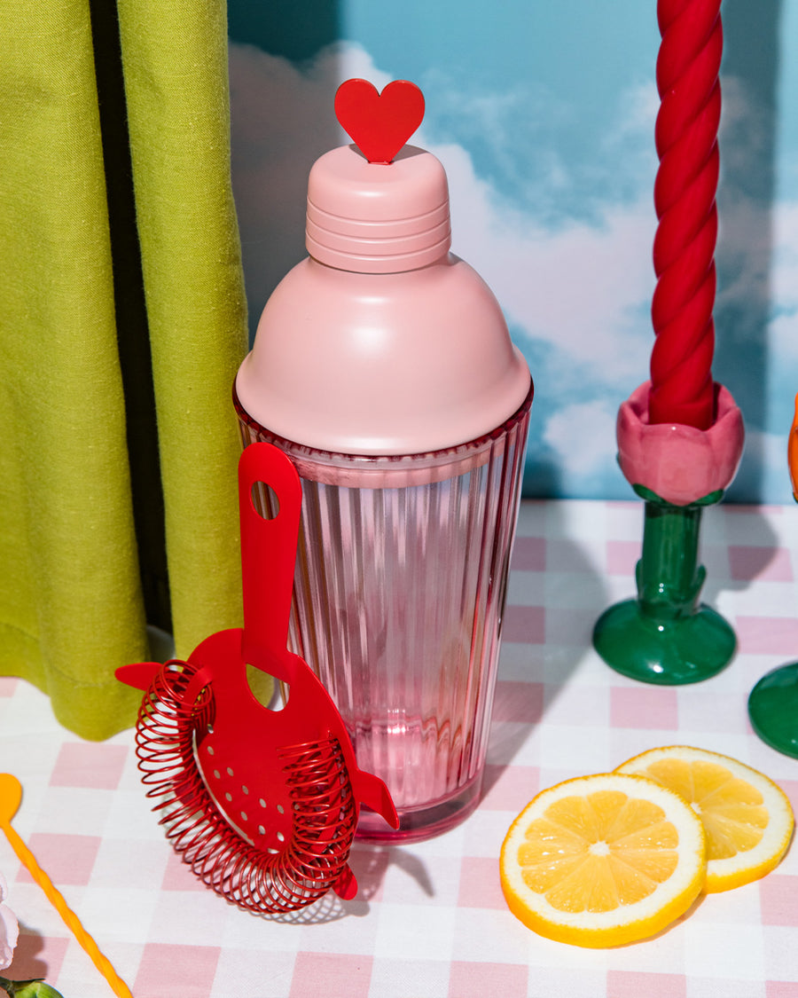 pink bar shaker with red heart top, red strainer, and yellow stirring stick on a pink checkered table