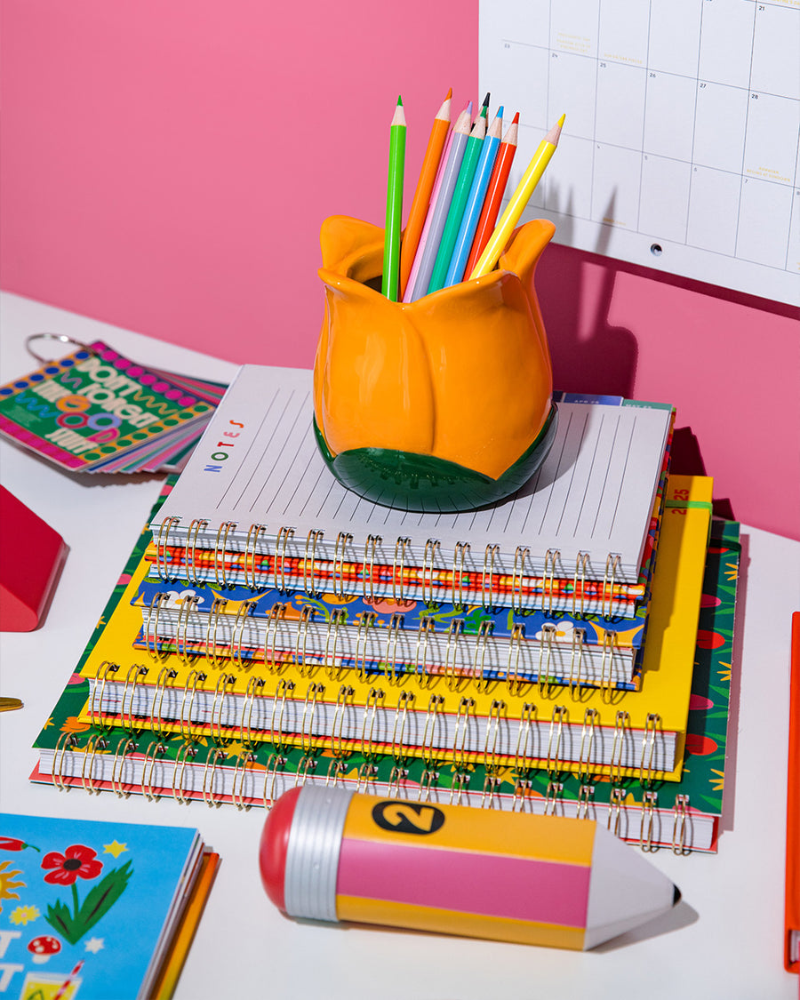 orange tulip shaped pencil holder with various pens inside on a stack of notebooks