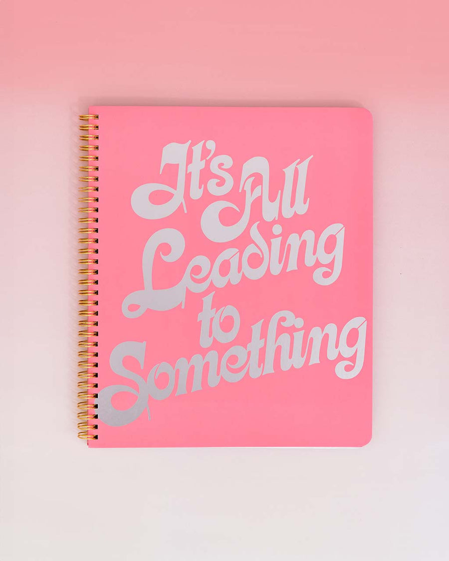 pink large notebook with white 'it's all leading to something' typography across the front