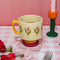 cream 10 oz ceramic mug with yellow handle, pink pedestal base and pink tulip print on a table next to a tulip candle holder