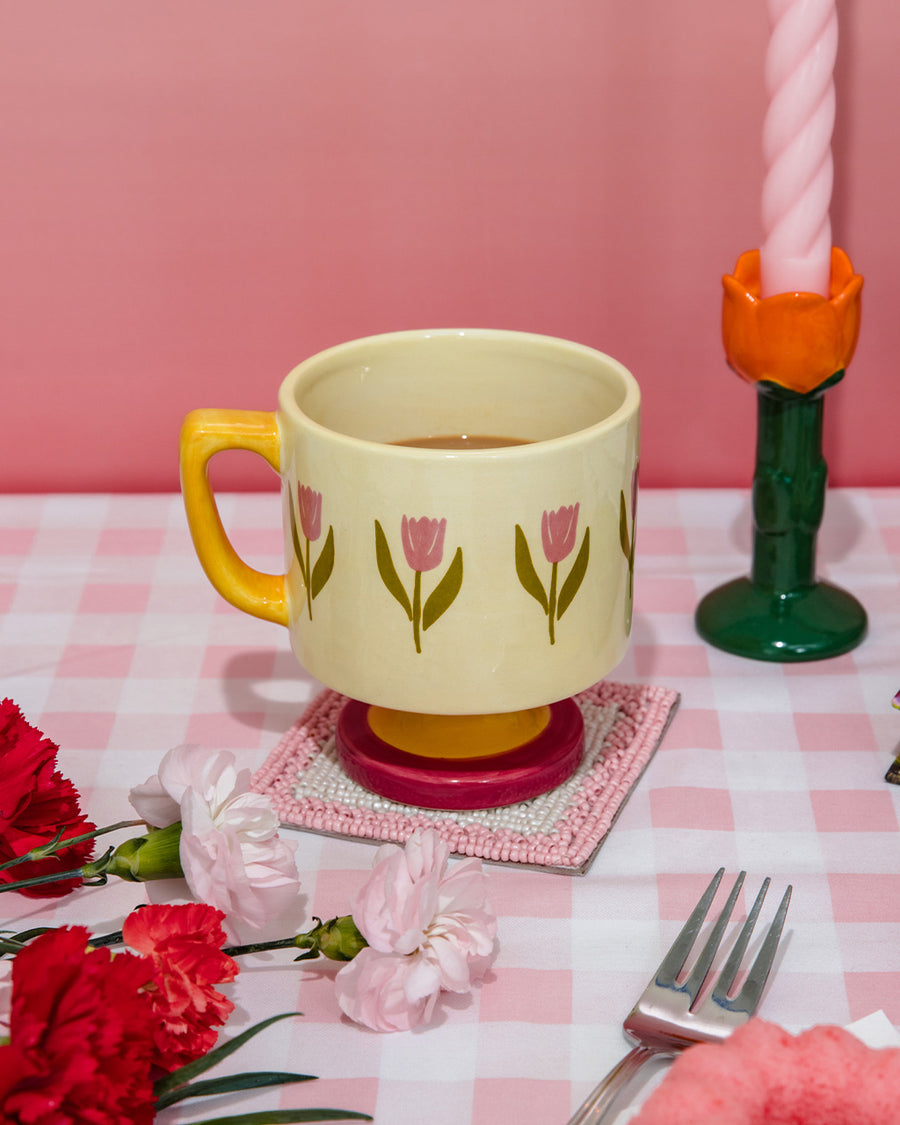 cream 10 oz ceramic mug with yellow handle, pink pedestal base and pink tulip print on a table next to a tulip candle holder