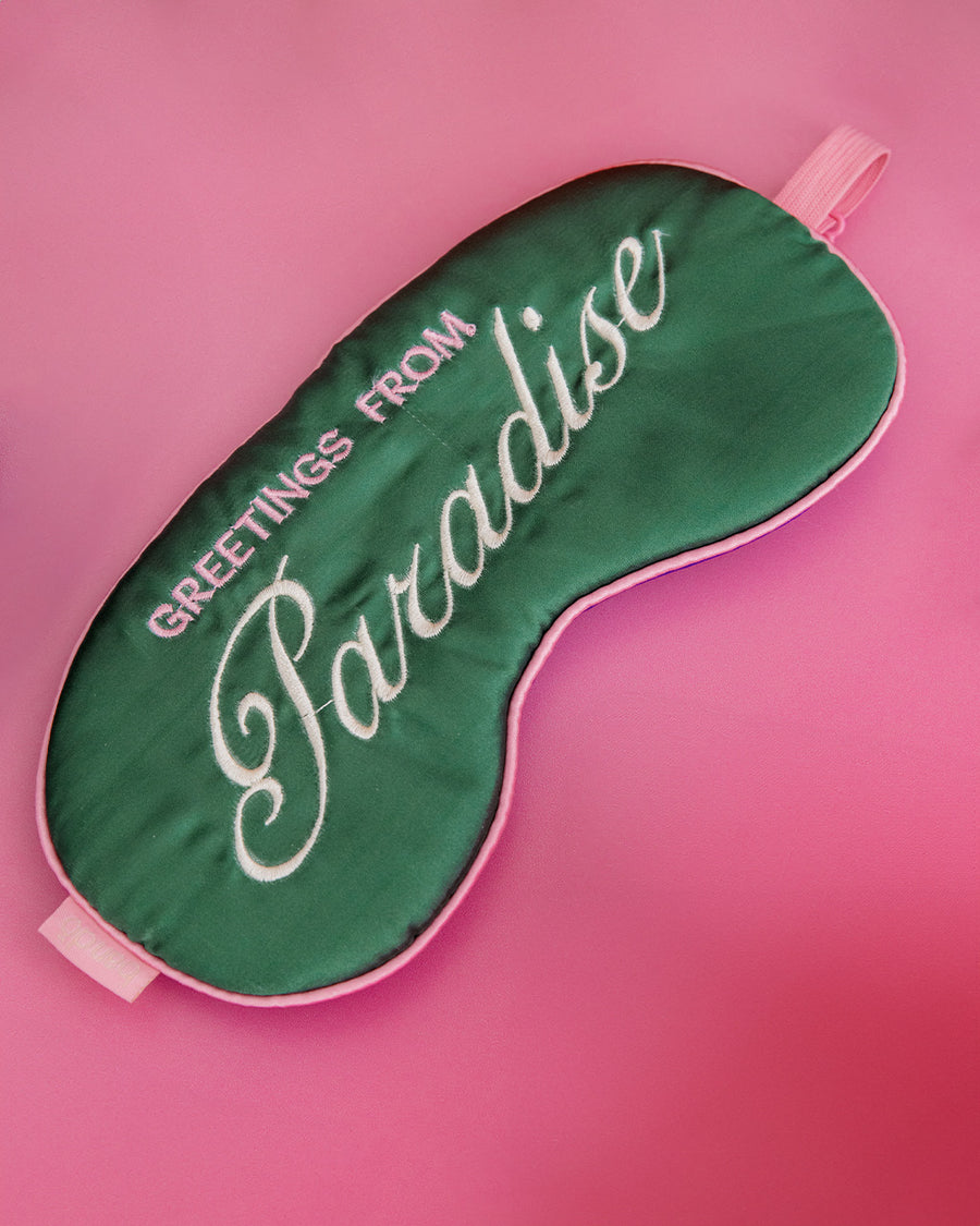 dark green sleep mask with pink and white embroidered 'greeting from paradise' with pink adjustable strap on a pink ground