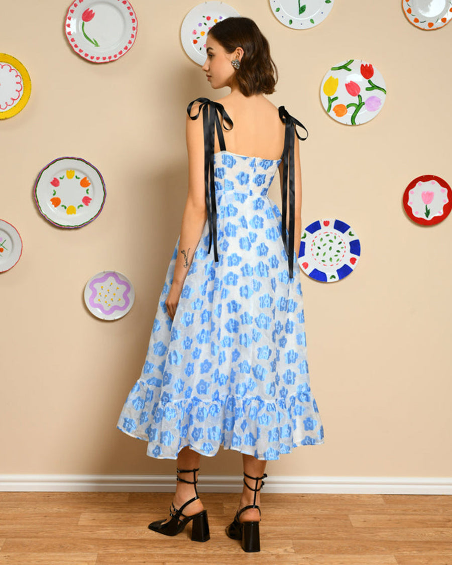 back view of model wearing white/light blue midi dress with ruffle hem, black tie straps and all over blue flower print