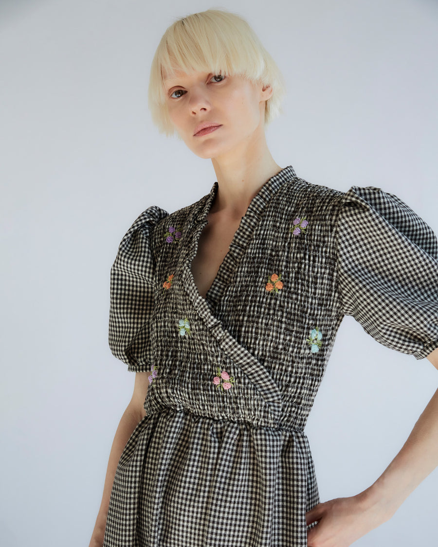 image of model with hand on hip in black and white smocked gingham dress with puff sleeves, faux wrap and colorful embroidered flowers on bust