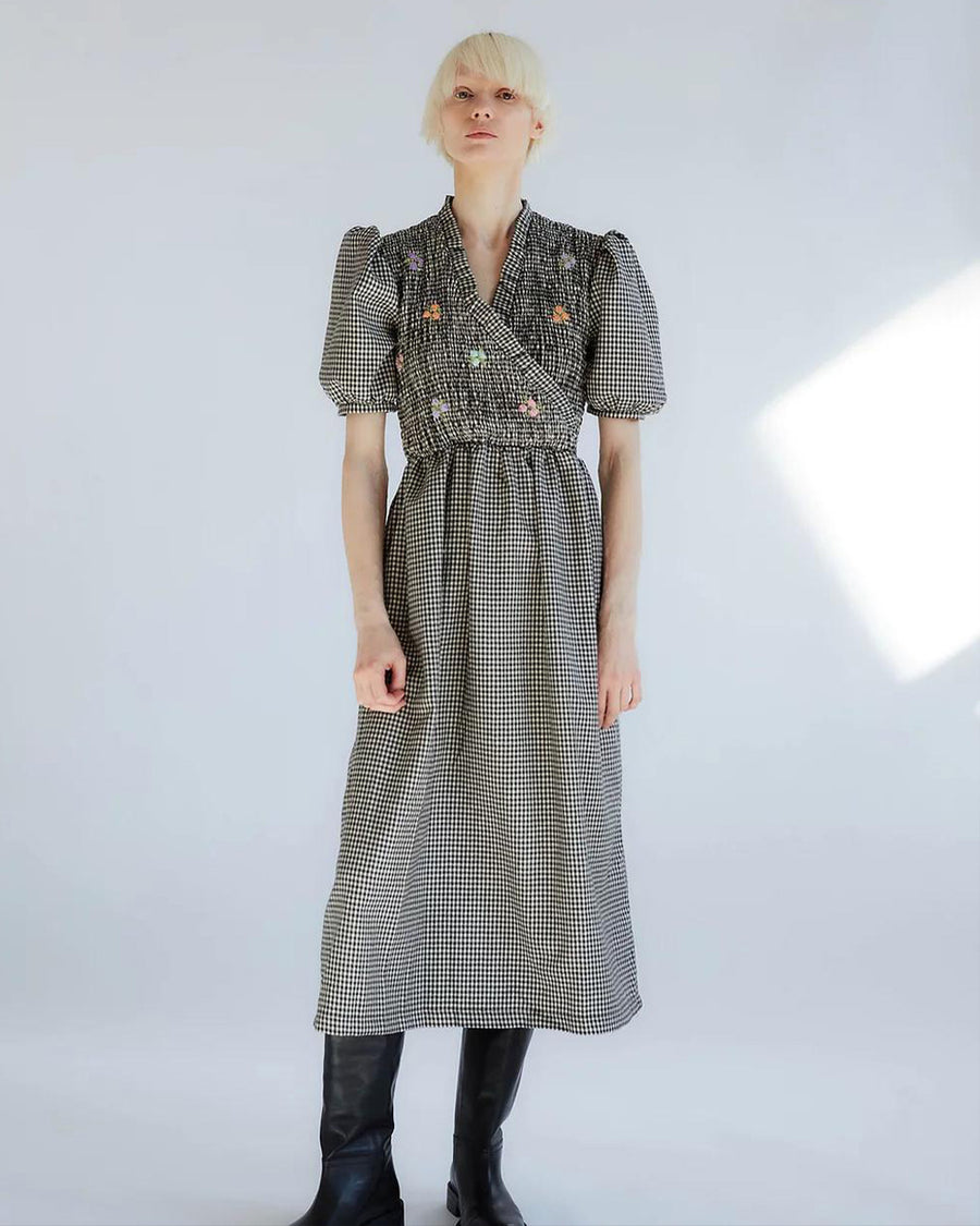 model wearing black and white smocked gingham dress with puff sleeves, faux wrap and colorful embroidered flowers on bust and black high boots