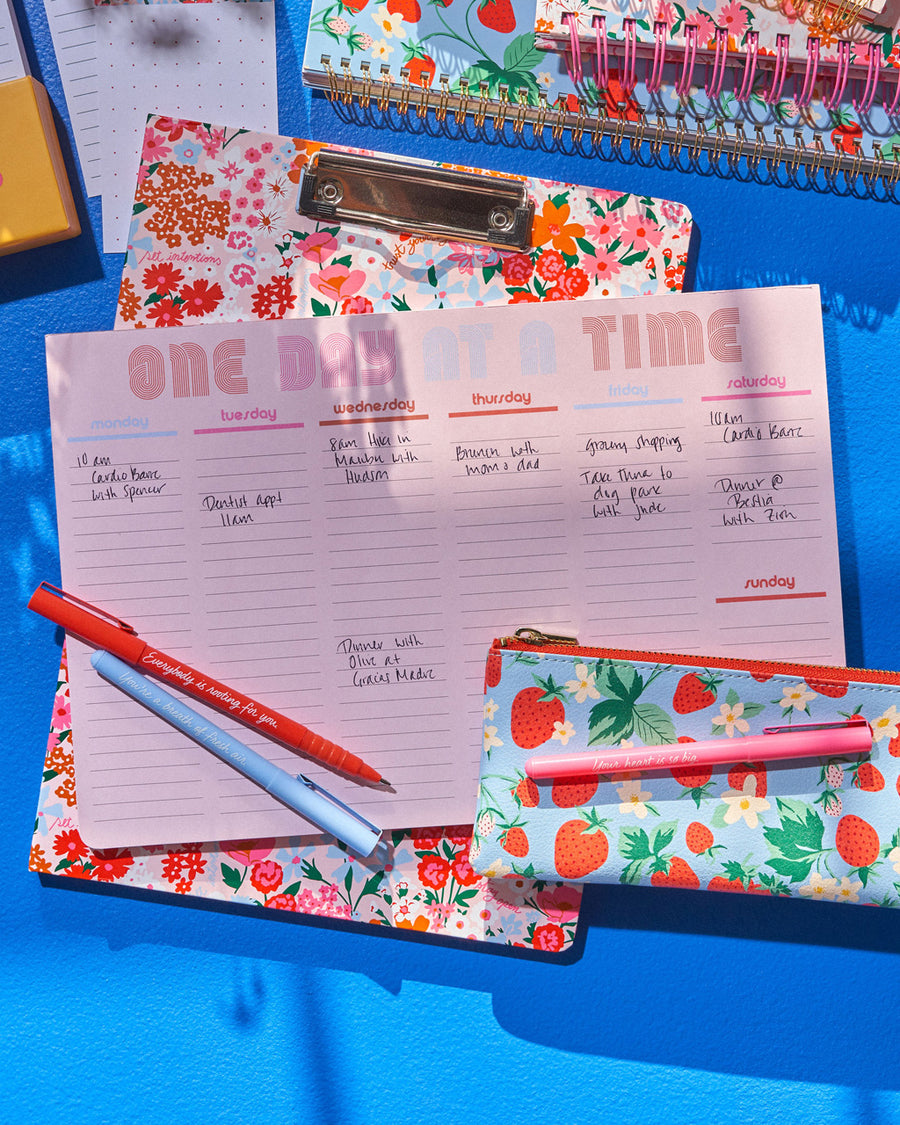 weekly deskpad in pink with "ONE DAY AT A TIME" text graphic at the top with pencil pouch and pens against blue backdrop shown with other desk accessories